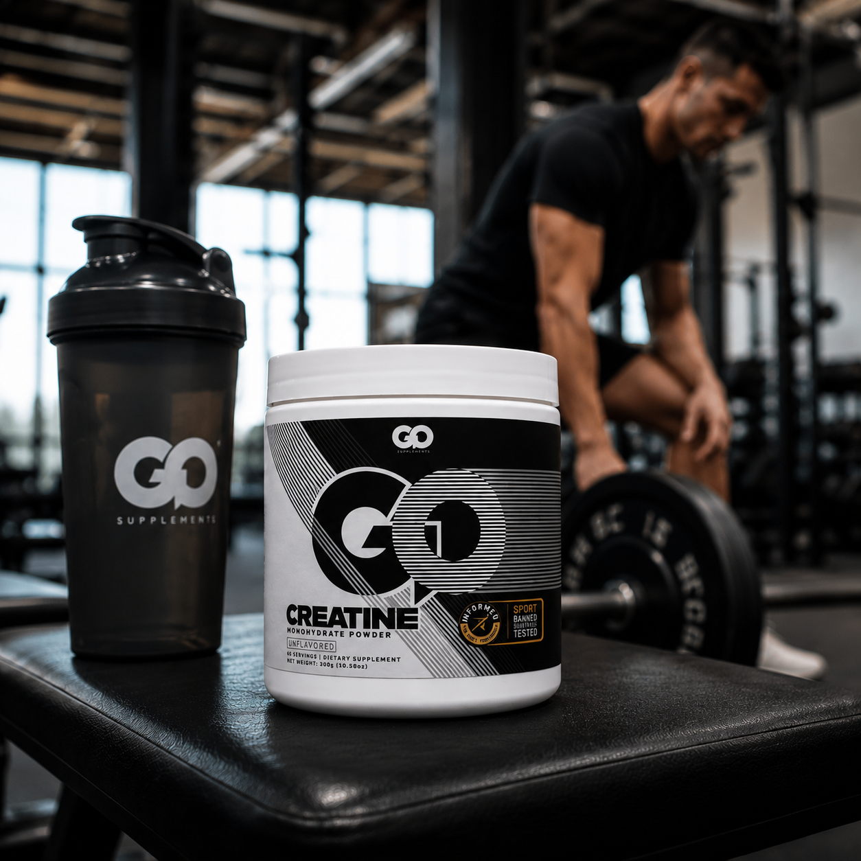 A white tub of GO Creatine powder and a black shaker bottle with a GO Supplements logo are placed on a gym bench, with a blurred man lifting weights in the background.